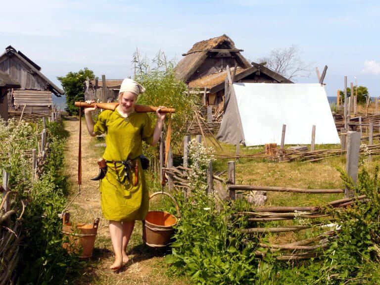 A young woman in Viking-age dress carrying two buckets on a wooden yoke through the reconstructed town at Foteviken, Sweden