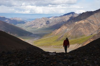 A hiker in a red jacket on a rocky ridge above a wide mountain valley in Denali National Park, Alaska